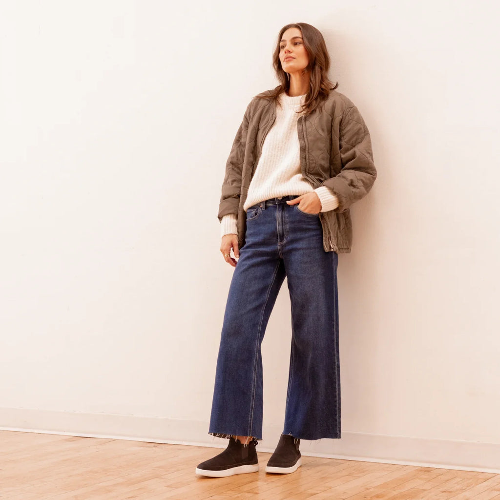 Woman standing wearing Neptune Athena black slip-on shoes facing left, styled with casual denim and jacket.