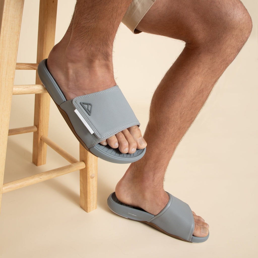Phantom Slab men's grey slide sandals, left foot facing forward close-up, right foot resting on stool leg, comfort design.