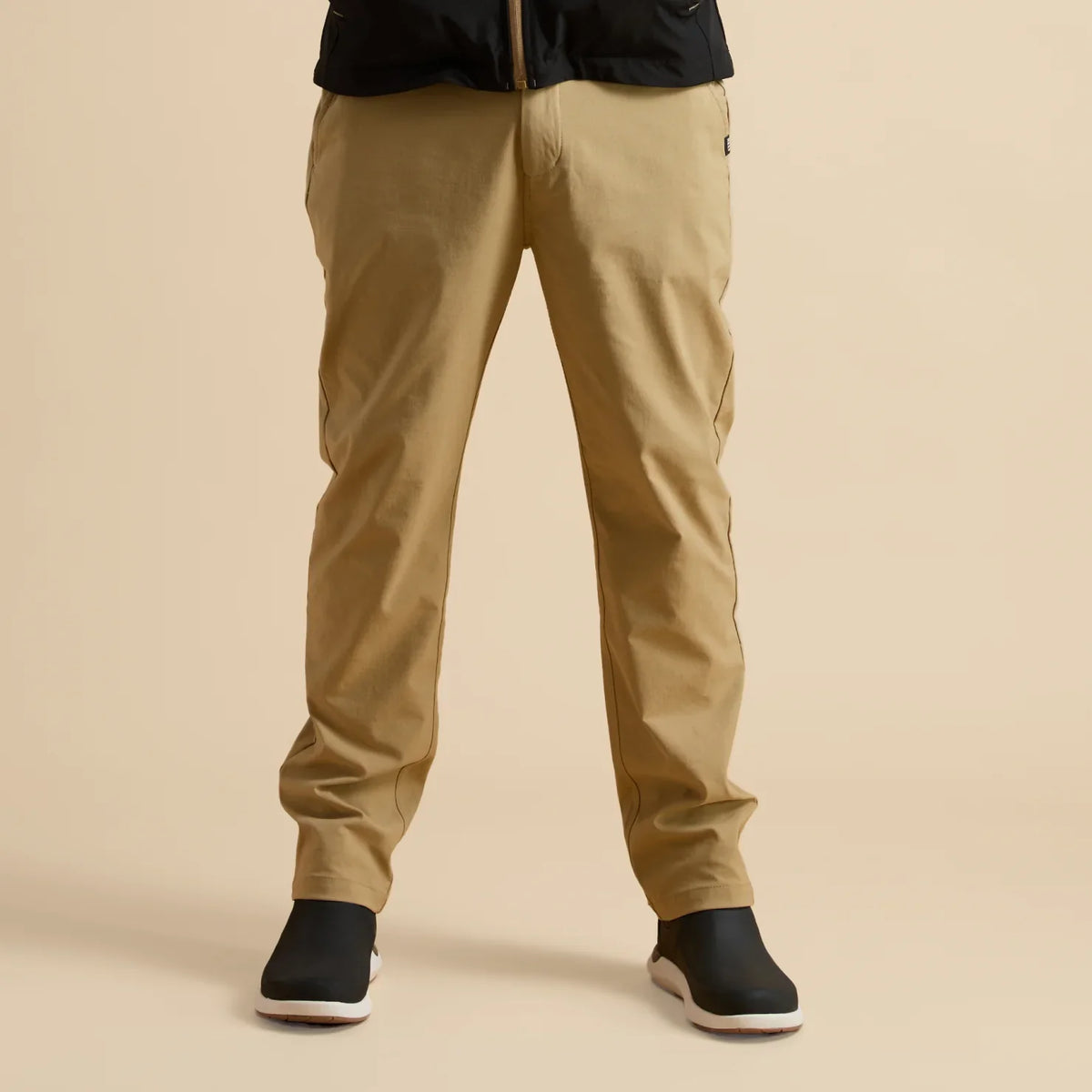 Front view of men's Carrick Stretch Pant in beige paired with black and white REEF slip-on shoes, beach-inspired comfort.