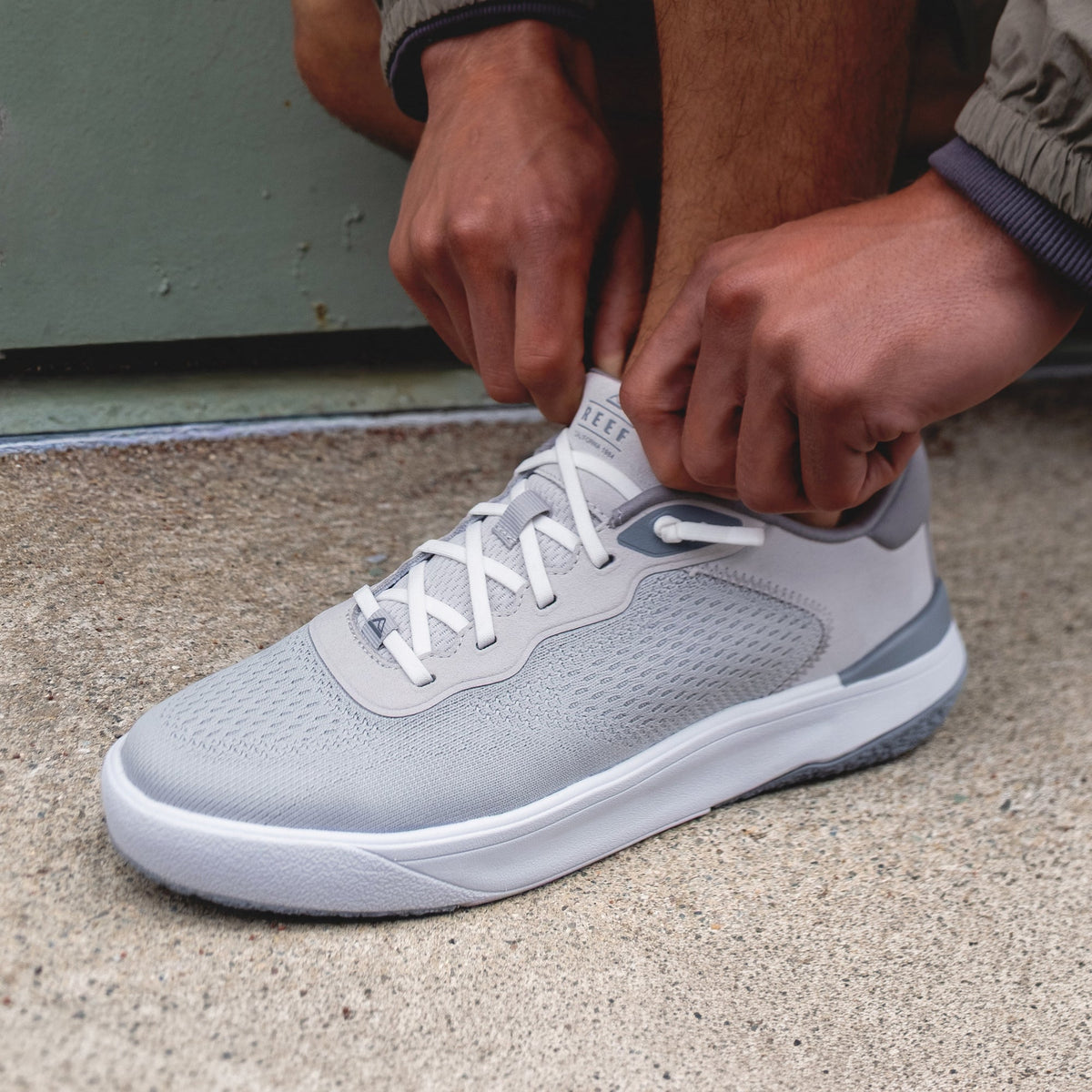 Shoreline Del Mar sneaker facing left, light grey breathable upper, cushioned sole, elastic lace detail, hands adjusting laces