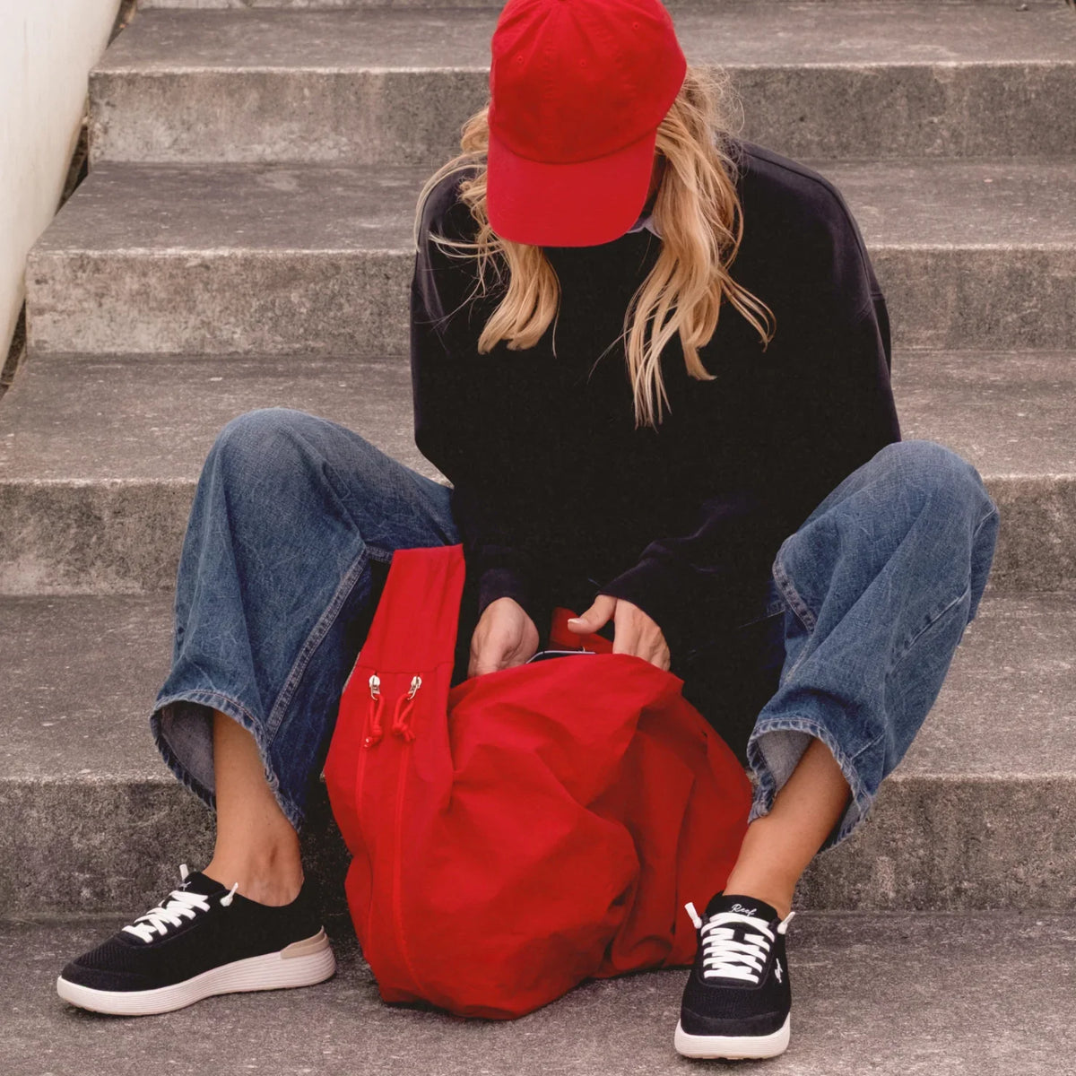 Woman wearing Shoreline Palisades sneakers facing forward, sitting on concrete steps with a red backpack and cap.