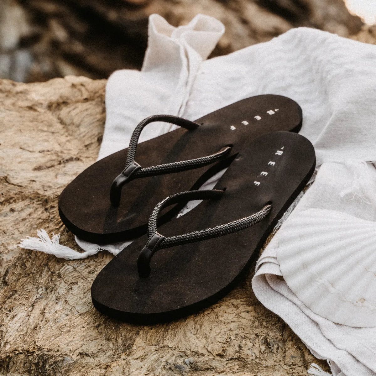 Bliss Bling Fling women's black flip flops facing left, close-up of top and straps on a beach towel, water friendly design.