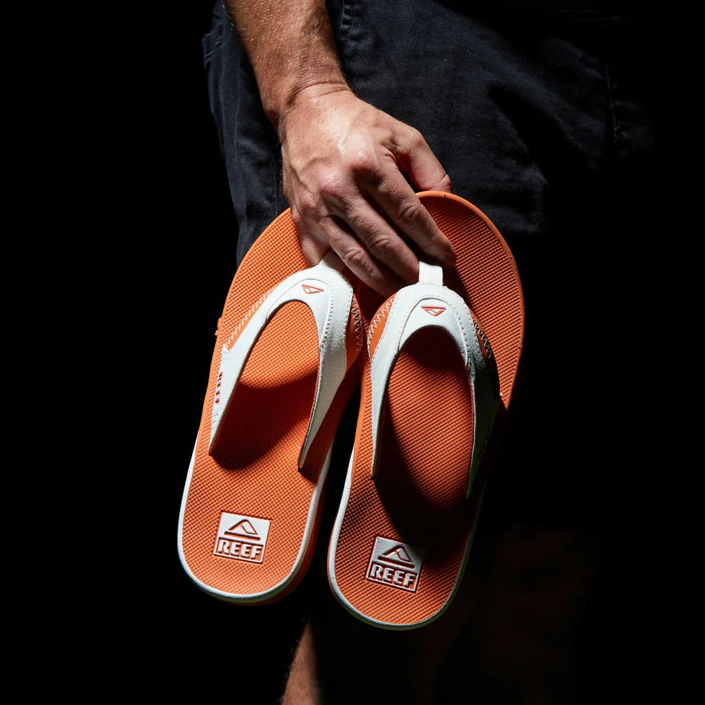 Hand holding a pair of men's Fanning Pregame flip flops facing sole up, showing orange footbed and white straps with arch support.