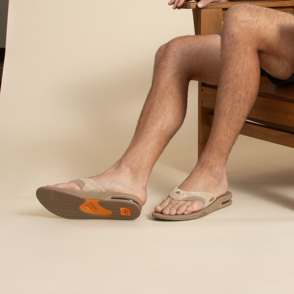 Fanning men's tan flip flop facing left with close-up on footbed and soft-rebound cushioning, surf-inspired comfort.