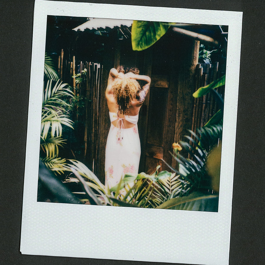 Woman wearing Belladona Dress facing away surrounded by tropical plants in a relaxed outdoor setting